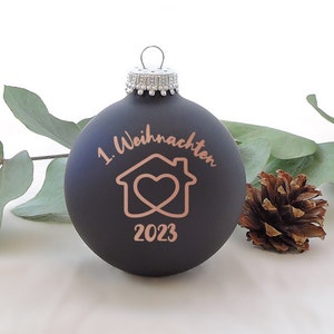 Könnte beinhalten: Eine mattschwarze Weihnachtskugel mit der Aufschrift "1. Weihnachten" und einem hausförmigen Design mit einem Herzen sowie der Jahreszahl 2023 in Kupferfarbe. Die Kugel hat eine silberne Kappe und wird mit Eukalyptusblättern und einem Tannenzapfen präsentiert.