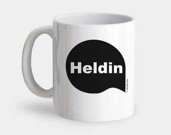 May include: White ceramic mug with a black speech bubble that says "Heldin" in white text.