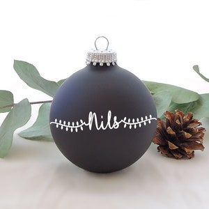 Könnte beinhalten: Mattschwarzer Weihnachtsschmuck mit dem Namen "Nils" in weißer Schrift, umrahmt von einer Lichterkette. Der Schmuck hat eine silberne Kappe und wird mit Grün und einem Tannenzapfen präsentiert.