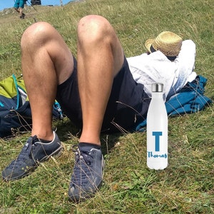 May include: White water bottle with a silver cap, featuring a large blue letter "T" and the name "Thomas" in a script font. The bottle is shown outdoors on a grassy hill.