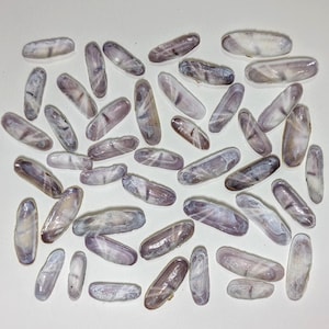 May include: An overhead shot displays numerous elongated seashells. The shells are primarily a pale lavender color, with subtle variations in tone and pattern. Some shells have darker streaks and markings, adding visual interest. The arrangement is scattered, suggesting a natural collection of beach finds.