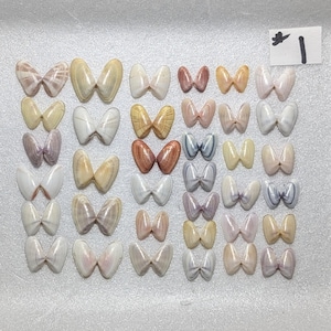 Könnte beinhalten: Eine Ansammlung von Muscheln, die in einem Raster angeordnet sind. Die Muscheln sind in verschiedenen Farben gehalten, darunter Weiß, Beige, Braun und Rosa. Jede Muschel hat die Form einer Fliege. Ein kleines weißes Etikett mit der Nummer "1" ist zu sehen.
