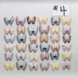 May include: A collection of thirty-six butterfly-shaped arrangements made from pairs of small, smooth bivalve seashells in various shades of cream, tan, grey, and lavender. A white paper tag with the handwritten black text #4 is at the top right.