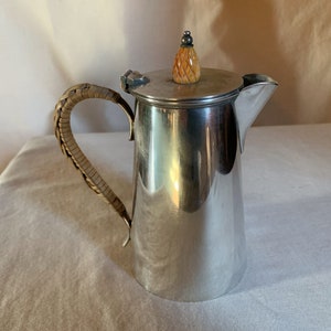 May include: A silver pitcher with a woven handle and a lid topped with a carved pineapple finial. The pitcher has a spout for pouring and is likely used for serving beverages.
