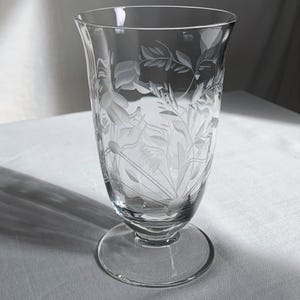 Könnte beinhalten: Klares Trinkglas mit geätztem Blumendesign. Das Glas hat einen ausgestellten oberen Rand, einen runden Fuß und einen Stiel. Das geätzte Design zeigt Blumen und Blätter. Das Glas wird wahrscheinlich für Wasser oder andere Getränke verwendet.