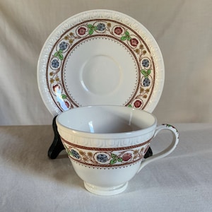 May include: A white teacup and saucer set. The cup and saucer have a decorative floral and rope design in red, blue, and green. The rim of the saucer has a repeating pattern. The cup has a handle.