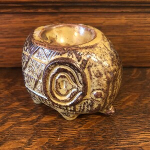 May include: A ceramic egg cup shaped like an elephant with brown and yellow glaze. The elephant has a detailed design on its body and a small hole on top for holding an egg.