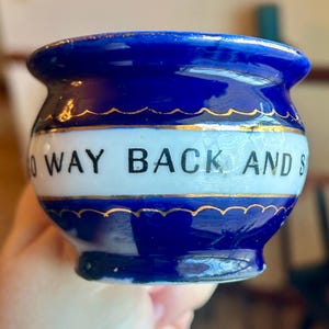 May include: A small, blue ceramic container with a white band featuring the text "NO WAY BACK AND S". The container has a gold trim and a rounded shape, held in a hand.