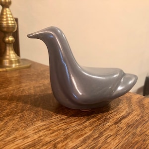 May include: A grey ceramic bird figurine with a smooth, glossy finish. The bird is perched on a wooden surface.