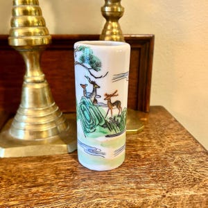 May include: A white ceramic vase with a rectangular shape. The vase features a hand-painted landscape with deer, trees, and mountains in shades of green, brown, and blue. The vase is approximately 4 inches tall.