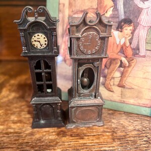 May include: Two miniature grandfather clocks with detailed designs. The clocks are dark brown with clock faces showing the time. One clock has a light-colored face, while the other has a darker face. The clocks are placed in front of a vintage illustration.