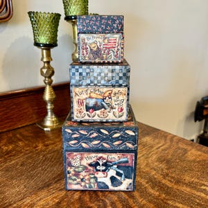 May include: Four stacked wooden boxes with colorful painted designs. The top box says "Be Proud" and features a frog. The second box says "A bit of caring goes a long way" and features a cat. The third box has a black and white checkered pattern. The bottom box says "Have fun." and features a cow.