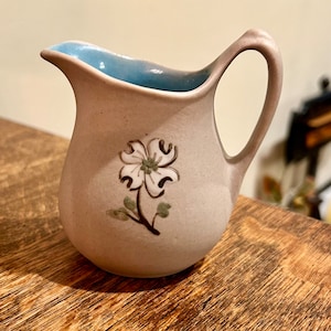 May include: A small, light brown ceramic creamer with a blue rim and a white flower design. The flower has a green stem and leaves.