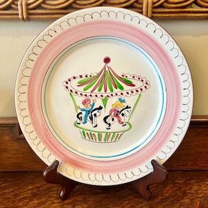 May include: A vintage ceramic plate with a pink rim and a white center featuring a hand-painted carousel with two children riding horses. The carousel is decorated with green, red, and white stripes.