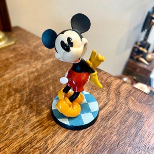 May include: A Mickey Mouse figurine featuring a black head with large round ears, a cream-coloured face, a black torso, red shorts, and yellow shoes. The character is posed on a circular base with a light and dark blue checkered pattern.