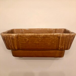 May include: A brown ceramic planter with a rectangular shape and a slightly raised lip. The planter has a textured surface and a slightly flared rim.