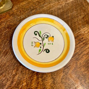 May include: A small, round ceramic plate with a white center and a yellow border. The center features a floral design with yellow flowers and green stems. The plate is likely used for serving small portions.