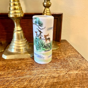May include: A small, white ceramic vase with a painted landscape scene featuring deer, trees, and mountains. The vase is cylindrical and has a glossy finish. The artwork includes green, blue, and brown colors.
