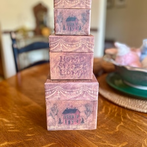 May include: A stack of four decorative boxes in varying sizes. The boxes are a muted reddish-purple with a distressed finish. Each box features a painted image of a house and the words "HOME SWEET HOME".