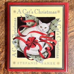 May include: A hardcover book titled "A Cat's Christmas" by Stefanie Samek. The cover features a white cat wrapped in red ribbon and a bow, set against a background of holly leaves. The book has a red border.