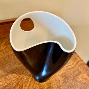 May include: A modern, two-tone ceramic vase. The top interior is white, contrasting with the glossy black exterior. It features a unique, asymmetrical design with a circular handle-like opening near the top.