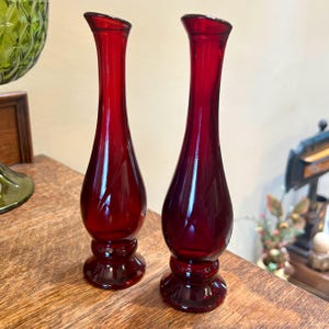 May include: Two red glass bud vases with a flared base and a narrow neck. The vases are on a wooden surface.