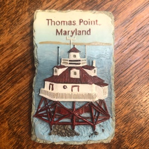 May include: A white and red lighthouse with a brown roof and a white railing. The lighthouse is on a stone base with the words "Thomas Point, Maryland" on the front.