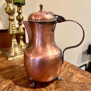 May include: A small copper teapot with a lid and a handle. The teapot has three feet and is decorated with a scalloped edge around the lid.