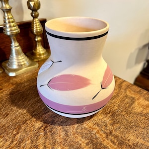 May include: A cream-colored ceramic vase with a wide opening and a rounded body. The vase features pink feather designs and black accents. A black band encircles the top, and a pink band with a black stripe is near the base. Decorative home decor.