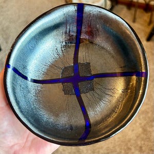 May include: A handmade, round ceramic bowl with a metallic silver and black finish. The bowl features a striking design of deep blue lines crossing the center. The bowl's interior has a textured surface, adding to its unique aesthetic.
