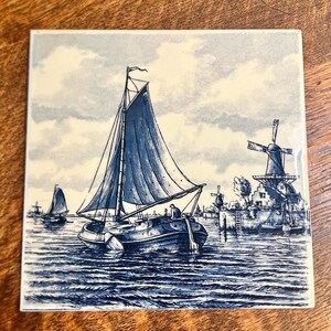 May include: A square ceramic tile featuring a blue and white illustration of a sailboat on the water. The scene includes a large central sailboat, a smaller boat in the distance, and a traditional windmill on the shore under a cloudy sky.