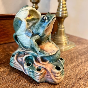 May include: A ceramic frog figurine in shades of green, brown, and cream. The frog is perched on a textured base with several holes. The figurine has a detailed, antique appearance, with a cracked glaze effect. Decorative item.