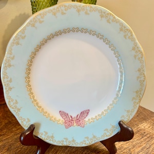 May include: A decorative plate with a light blue rim and a white center. The rim features a gold floral design and a gold heart-shaped border. A pink butterfly is in the center of the plate. The plate is on a wooden stand.