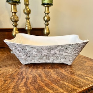 May include: A rectangular white and brown ceramic bowl with a textured basketweave pattern on the exterior. The bowl has a smooth white interior and is displayed on a wooden surface. Decorative brass candlesticks are in the background.