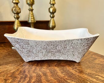 May include: A rectangular white and brown ceramic bowl with a textured basketweave pattern on the exterior. The bowl has a smooth white interior and is displayed on a wooden surface. Decorative brass candlesticks are in the background.