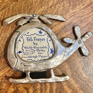 May include: A pewter helicopter-shaped picture frame with a cream-colored insert featuring blue stars and text. The frame has a propeller and tail rotor. The text reads "Folk Frames by Ruth Reynolds Darby for Shleigh Manor."
