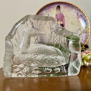 May include: A clear glass sculpture of a loon bird in a naturalistic setting. The bird is detailed with textured feathers and is surrounded by etched reeds and water plants. The sculpture is displayed in front of a decorative plate.