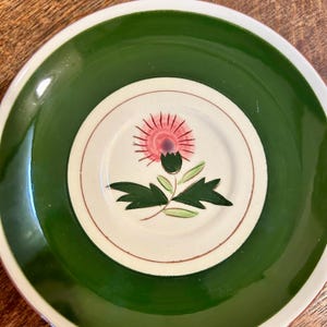 May include: A ceramic plate with a green border and a white center. The center features a pink and purple thistle flower design with green leaves. The plate has a white rim and is set against a wooden surface.