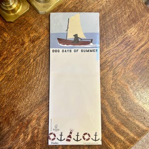May include: A rectangular notepad with a nautical theme. The top features a sailboat with a dog silhouette and the text "DOG DAYS OF SUMMER." The bottom has nautical icons. The notepad is on a wooden surface.