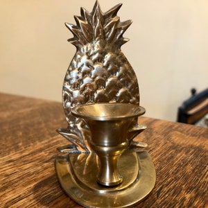 May include: A brass pineapple-shaped candle holder with a single candle cup. The holder is detailed with a textured pineapple design.