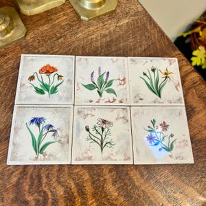 Puede incluir: Seis baldosas de cerámica cuadradas con diseños florales pintados a mano. Cada baldosa presenta una flor diferente en colores vibrantes como naranja, morado, azul y amarillo, sobre un fondo moteado blanquecino. Las baldosas están dispuestas sobre una superficie de madera.
