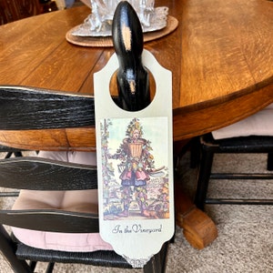 May include: A wooden door hanger with a vintage illustration of a person dressed in vines and holding a basket of grapes. The text "In the Vineyard" is printed at the bottom of the hanger.