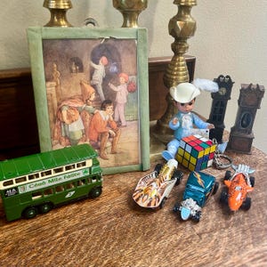 May include: A collection of vintage toys and decorative items on a wooden surface. Includes a green toy bus, a framed illustration, miniature clocks, a doll, and toy cars. The bus has the text "Céad Míle Fáilte".