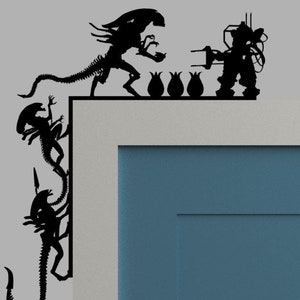 May include: Black and white silhouette wall decal featuring three alien creatures and a robot. The aliens are climbing a wall and the robot is holding a weapon. The decal is on a gray wall with a blue door.