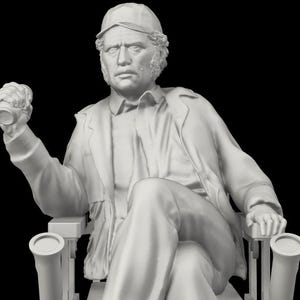 Op de afbeelding: Een gedetailleerde, monochrome sculptuur van een zittende figuur met een pet, jas en broek. De figuur houdt een klein blikje in zijn opgeheven hand. De sculptuur zit in een stoel met armleuningen.