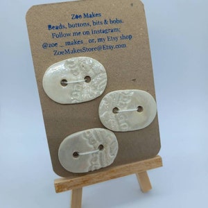 May include: Three cream-coloured, oval buttons with a textured surface are displayed on a small wooden easel. The buttons are attached to a brown card with the text "Zoe Makes" and contact information.