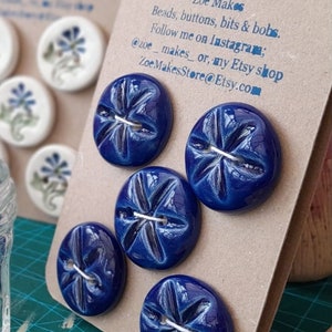 May include: Six blue ceramic buttons with a white star design. The buttons are displayed on a brown card with the text "Zoe Makes Beads, buttons, bits & bobs. Follow me on Instagram; @zoe_makes_ or, my Etsy shop ZoeMakesStore@Etsy.com"