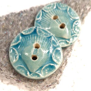 May include: Two round, turquoise ceramic buttons with a seashell design. Each button has two holes and a crackled glaze finish. The buttons are set against a textured, gray background.
