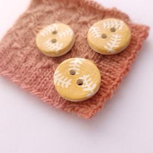 May include: Three yellow buttons with white leaf designs. The buttons are on a pink knitted fabric.