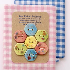 May include: A collection of colorful, hexagonal buttons with a speckled glaze, arranged on a cardboard tag. The tag reads "Zoe Makes Buttons." The buttons are set against pink and blue gingham fabric.